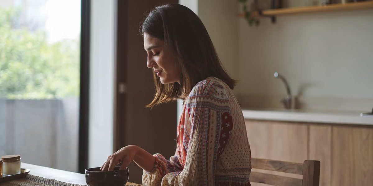 Woman in patterned dress enjoying breakfast at home near a sunlit window.