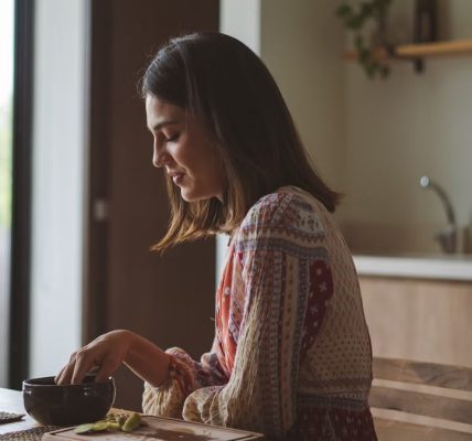 Woman in patterned dress enjoying breakfast at home near a sunlit window.