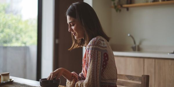 Woman in patterned dress enjoying breakfast at home near a sunlit window.
